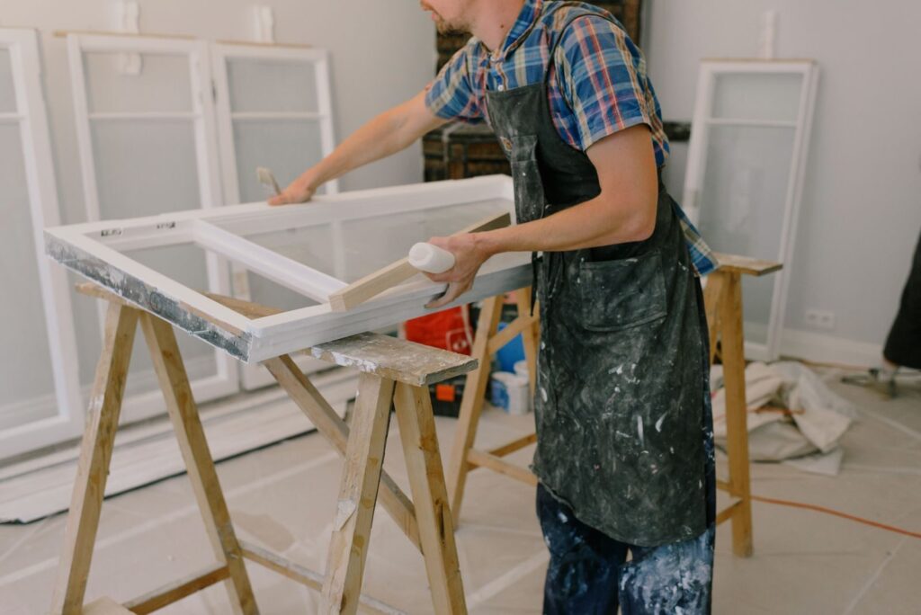 Unrecognizable male wearing dirty apron standing near window with white frames on wooden saw horse while working in professional studio