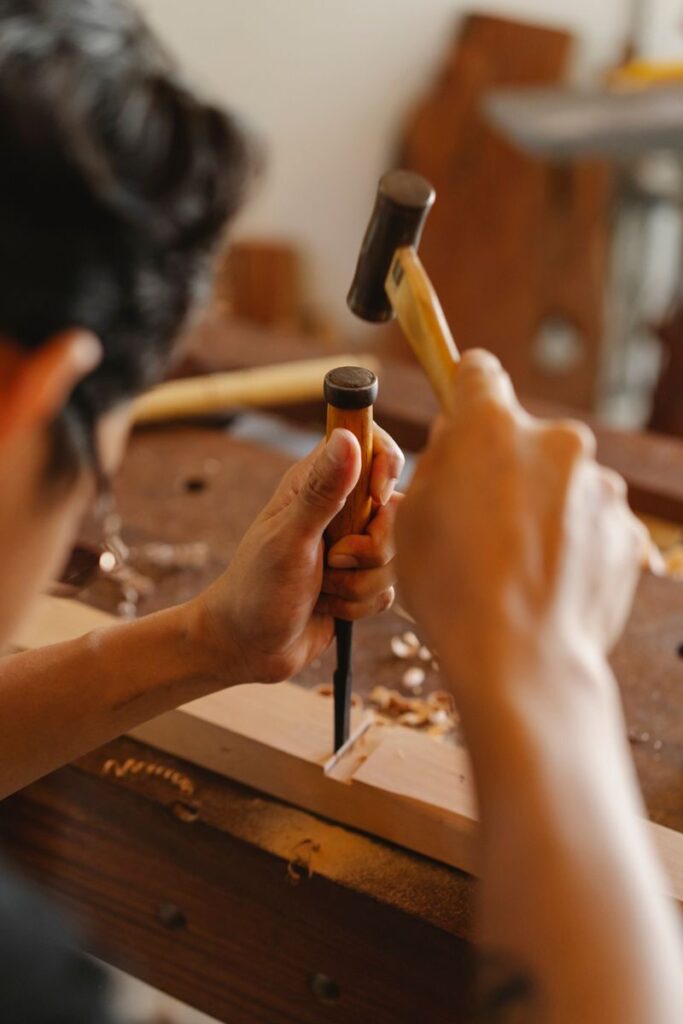 Anonymous carpenter in glasses forming wooden detail with hammer and chisel while working in joinery