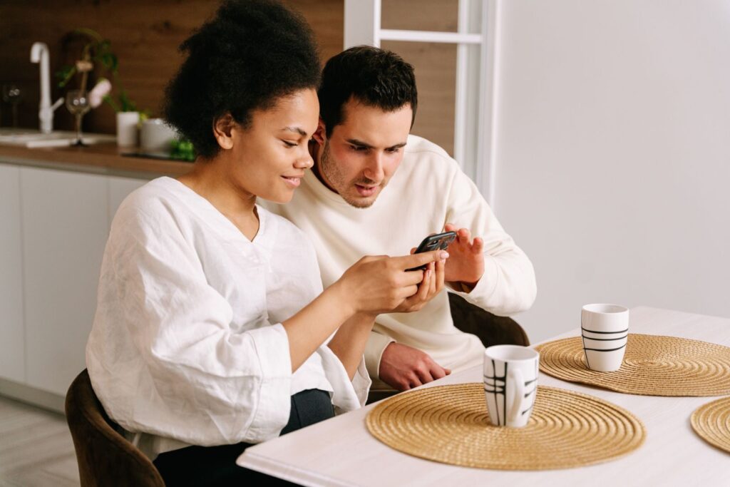 Happy couple sitting in kitchen browsing online shopping on smartphone, sharing moments together.