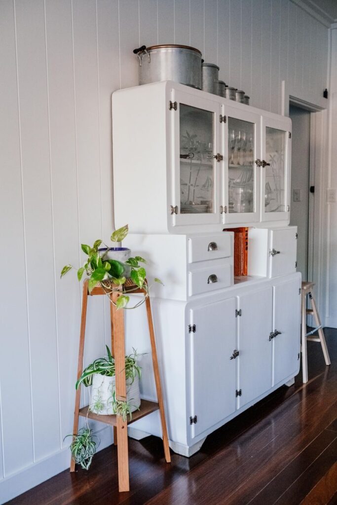Elegant white vintage restored painted kitchen cabinet with glass doors and potted plants in a serene home setting.