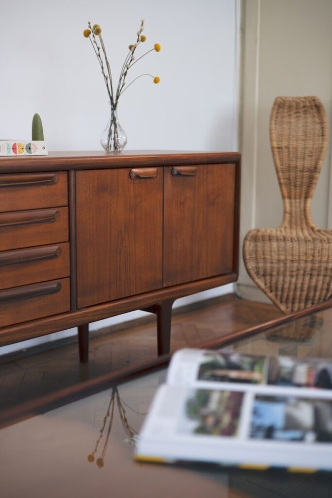 A vintage Nordic style wooden cabinet  from the 60s with a vase of flowers on top of it and a wicker chair