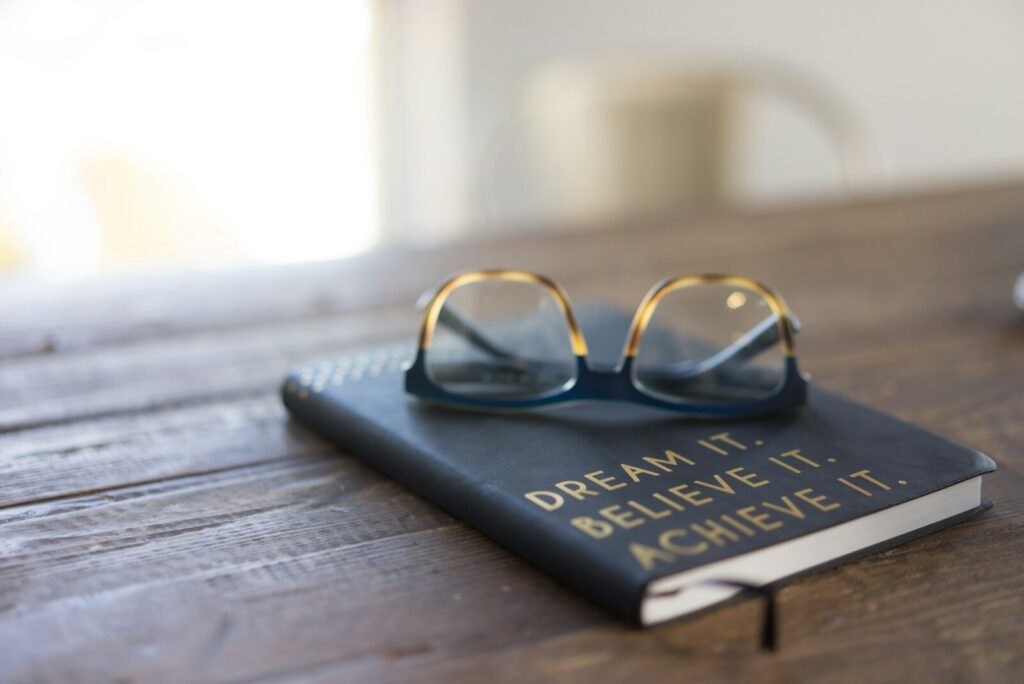 Black and brown eyeglasses on book on brown wooden table with a journal with an inspiration quote