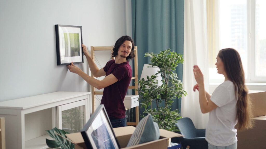 a man and a woman standing in a room with moving boxes, holding a picture up against the wall